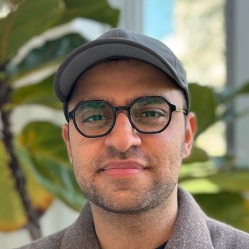 Moeed in a cap and round glasses, smiling slightly, beside a fiddle-leaf fig.